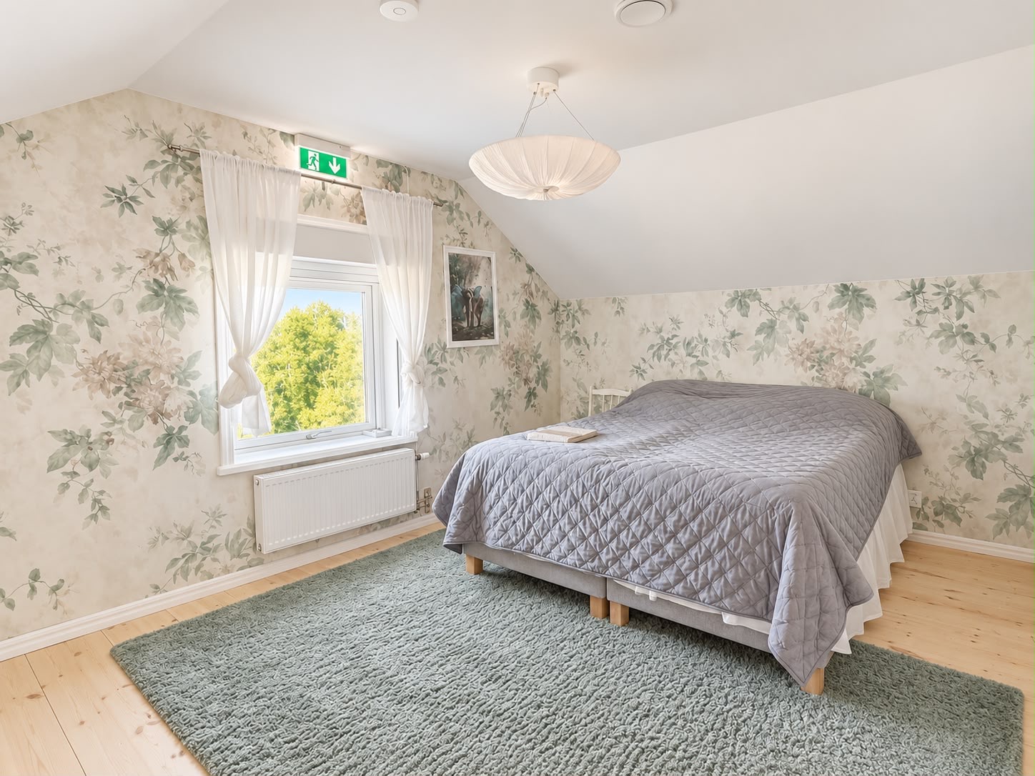 Ljust sovrum med blommig tapet, dubbelsäng och fönster mot grönskan